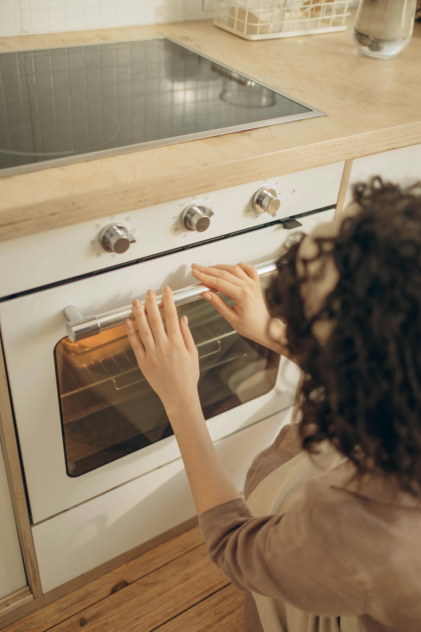 Une femme adulte aux cheveux bouclés utilisant un four à gaz moderne chez elle pour faire de la pâtisserie.