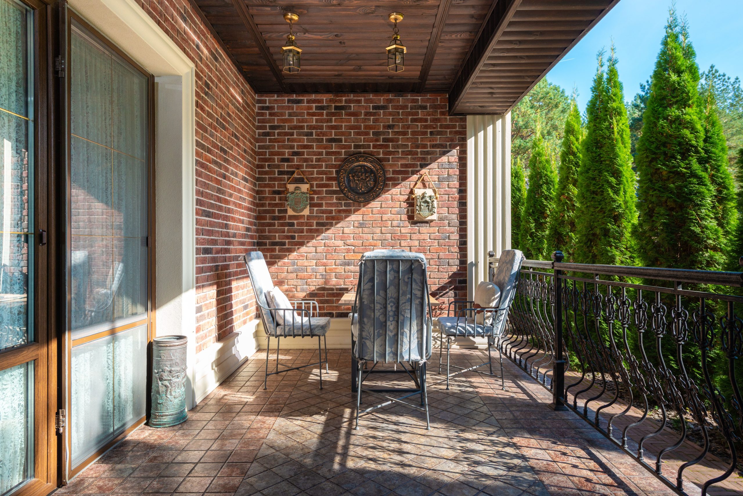 A cozy terrace with chairs and a brick wall under a clear, sunny sky featuring lush green trees. Une terrasse confortable avec des chaises et un mur en briques sous un ciel dégagé et ensoleillé avec des arbres verts luxuriants.
