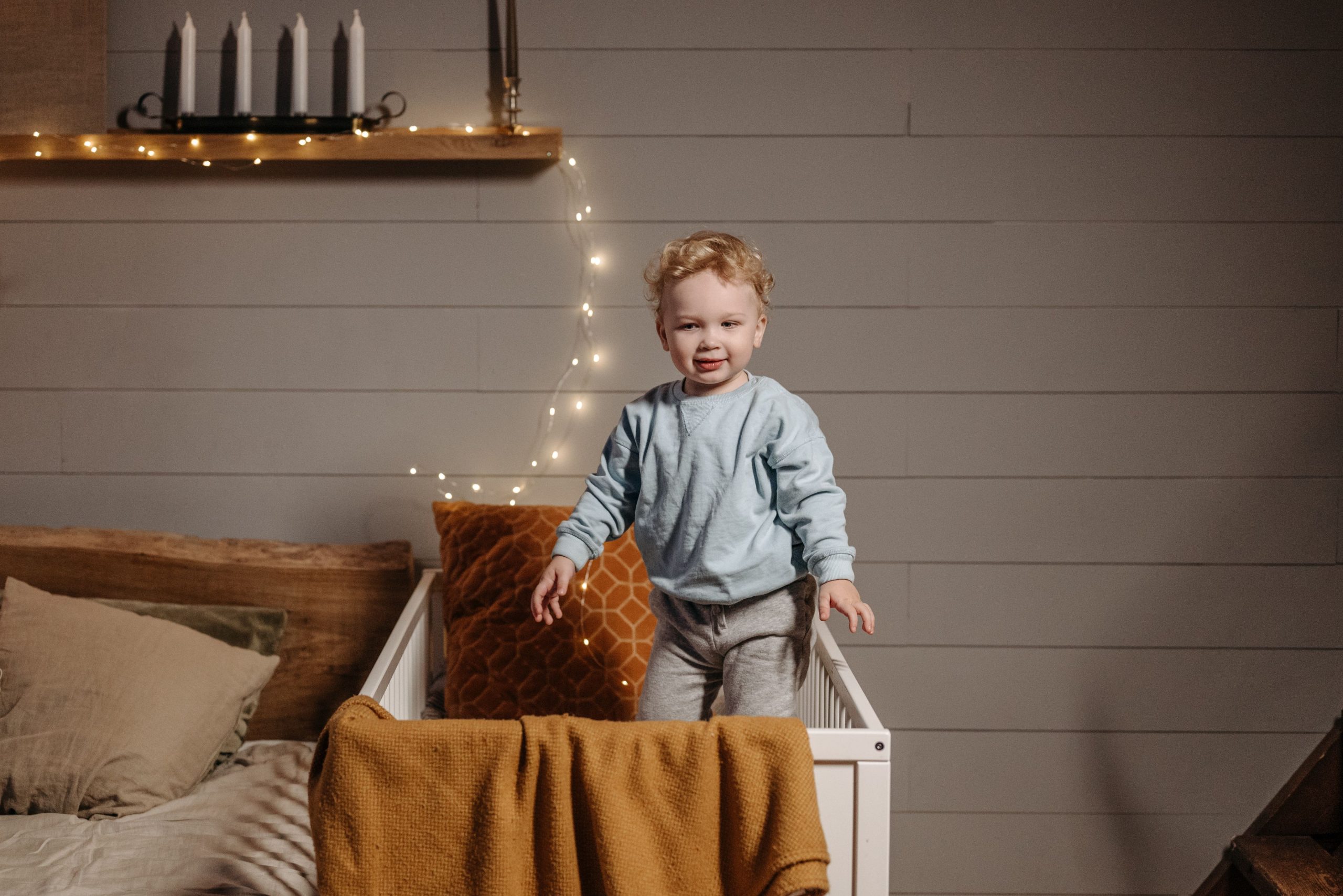 A cute baby standing in a crib with cozy decor and fairy lights in a bedroom. Un bébé mignon debout dans un berceau avec une décoration douillette et des guirlandes lumineuses dans une chambre.