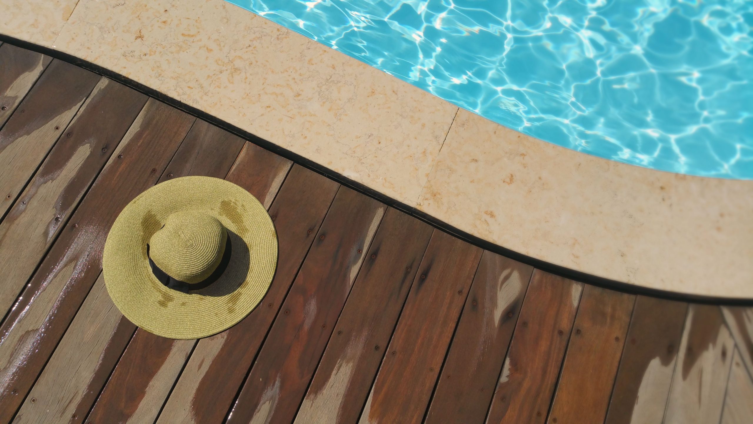 A straw hat rests on a wooden deck beside a sparkling blue swimming pool, perfect for summer relaxation. Un chapeau de paille repose sur une terrasse en bois à côté d'une piscine bleu étincelant, parfait pour la détente estivale.