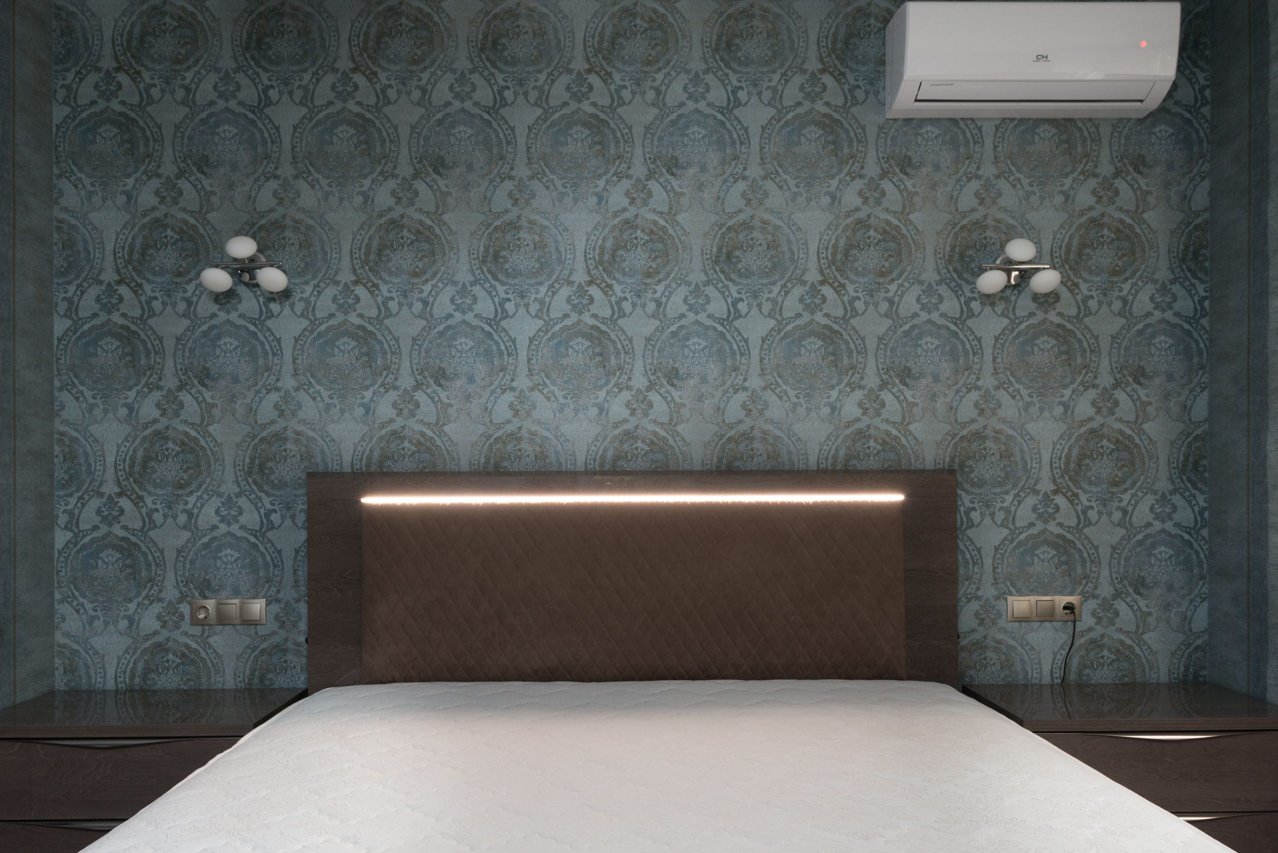 White mattress on bed with brown headboard placed between bedside tables at wall with ornamental wallpaper and decorative white sconces carpet head Matelas blanc sur lit avec tête de lit marron placée entre tables de nuit au mur avec papier peint ornemental et appliques décoratives blanches tapis tête